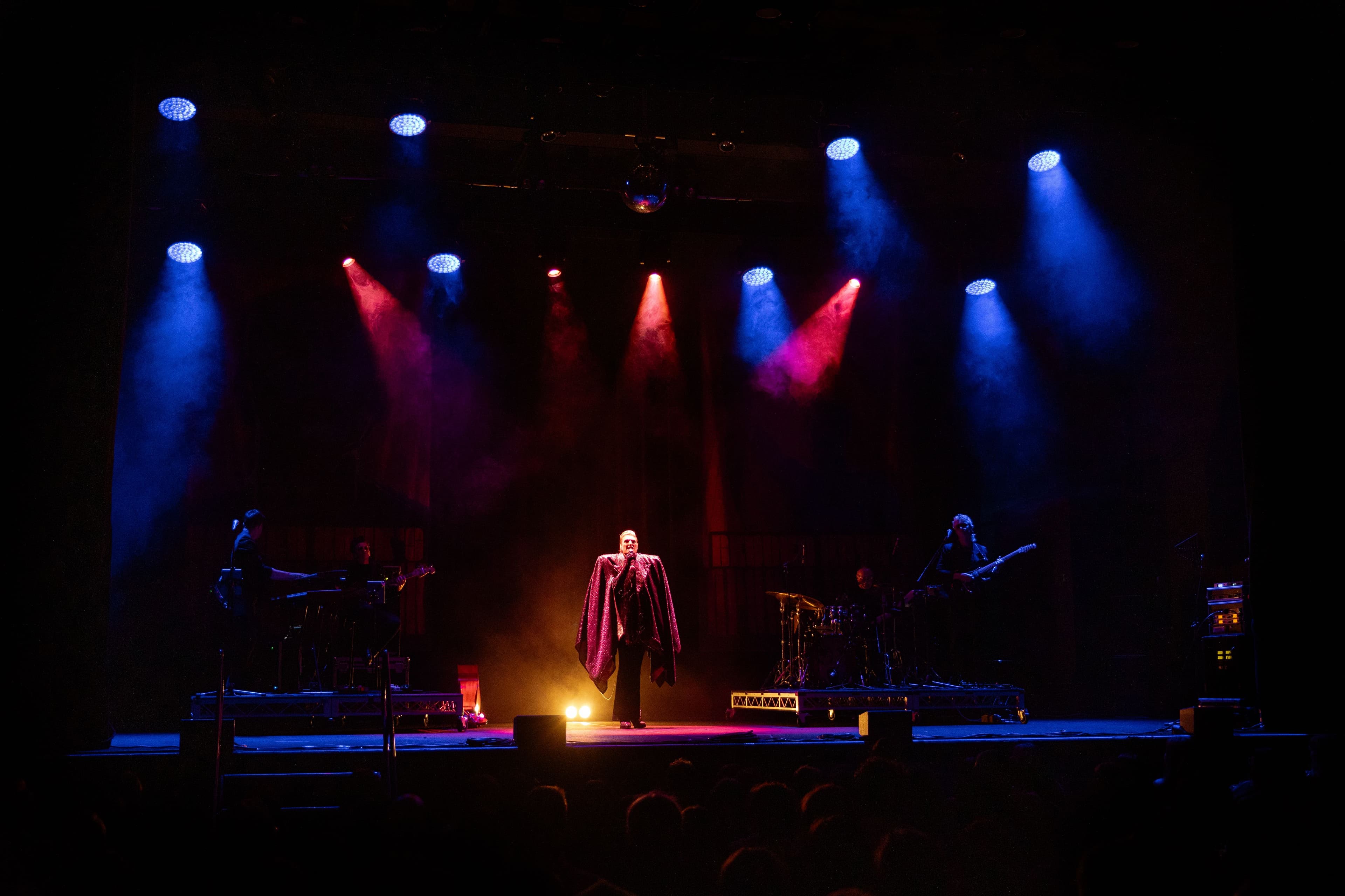 A performer in a cape stands center stage under dramatic red and blue lighting, with band members playing instruments on both sides.