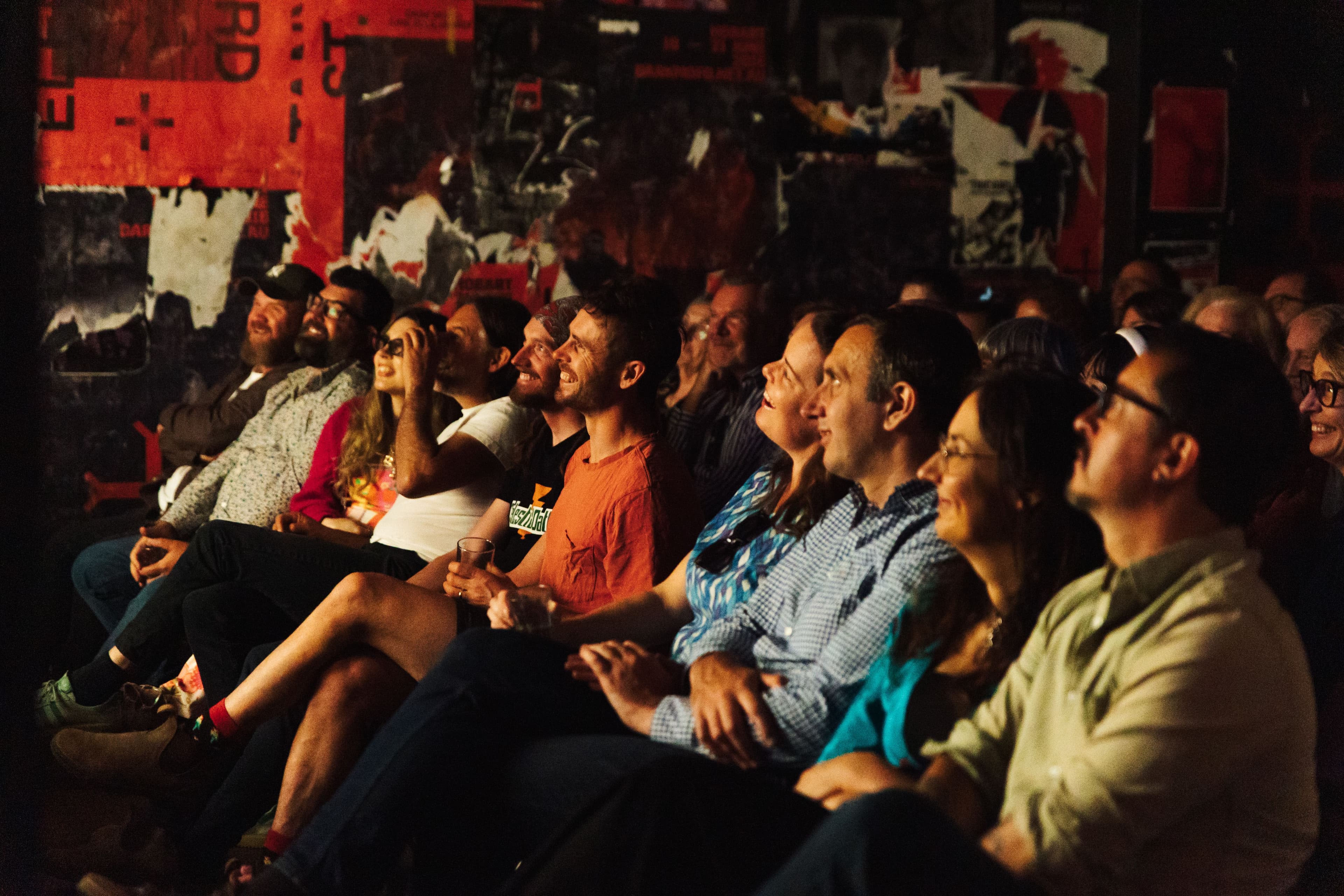 Audience members sit closely together in a dimly lit room, watching an event on stage and smiling. A red and black abstract mural is visible on the wall behind them.