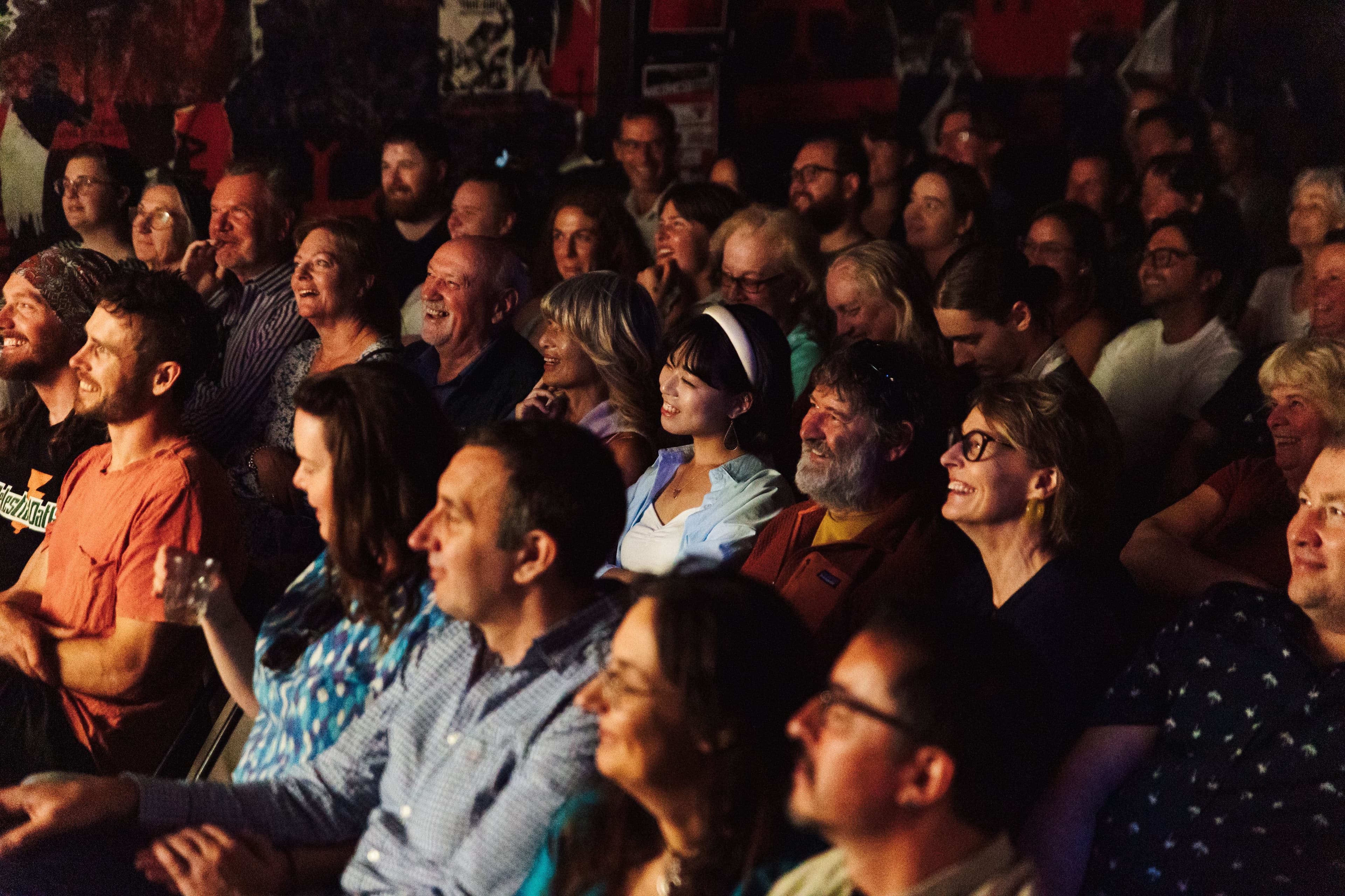 A smiling audience seated closely together, enjoying a live performance in a warmly lit venue.