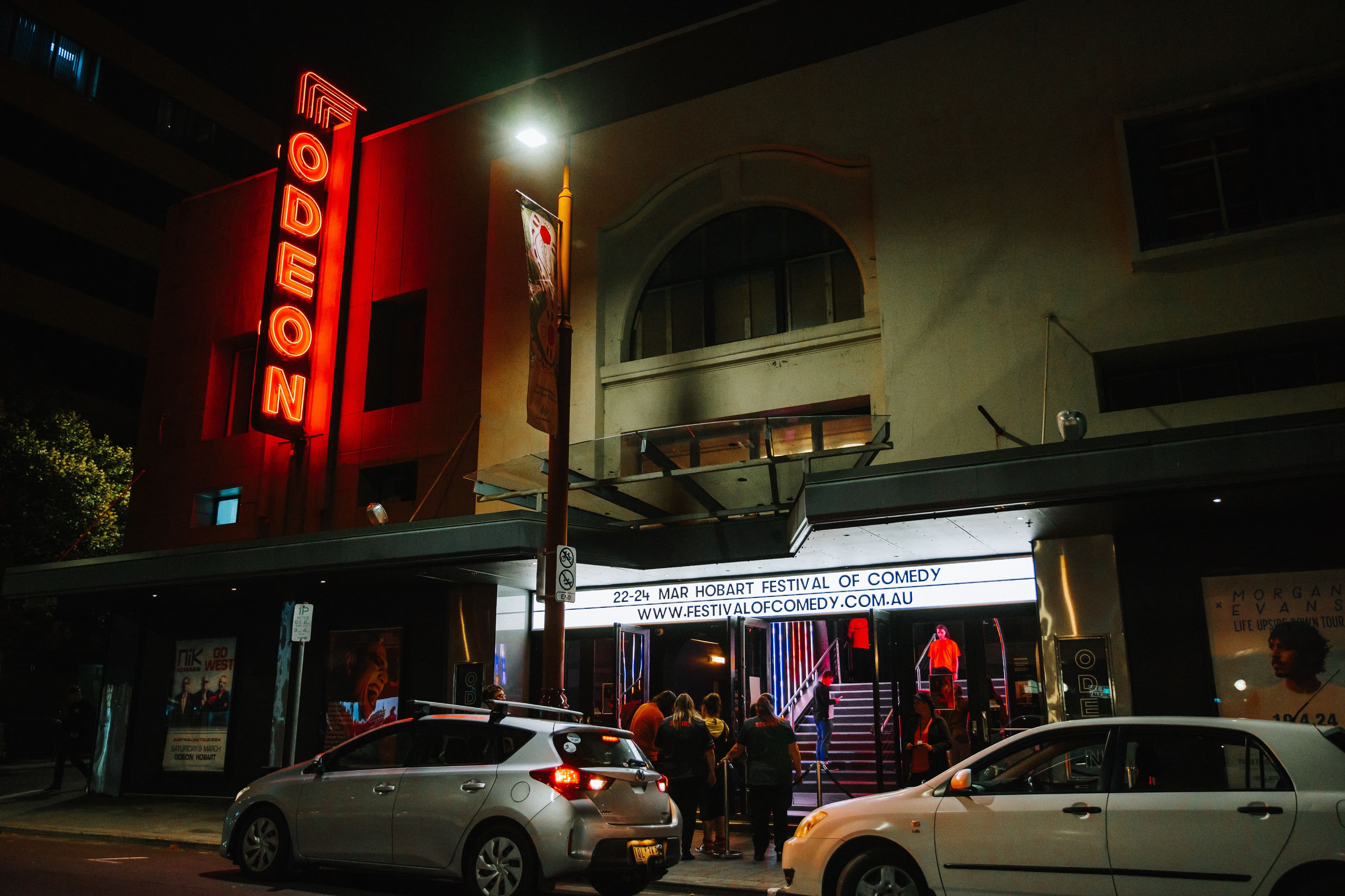 Street view of the Odeon Theatre at night, featuring a glowing neon sign, a comedy festival marquee, and people entering. Cars parked outside.