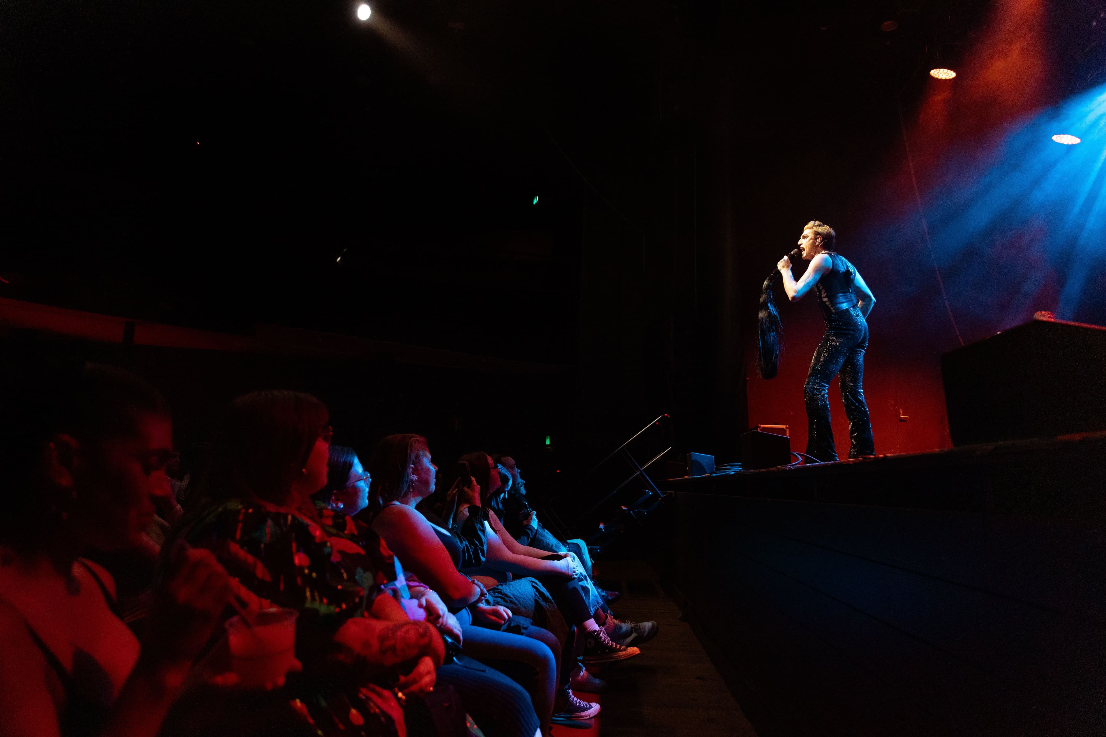 A performer in sequined pants sings on stage under red and blue lights as the audience watches closely from the front row.