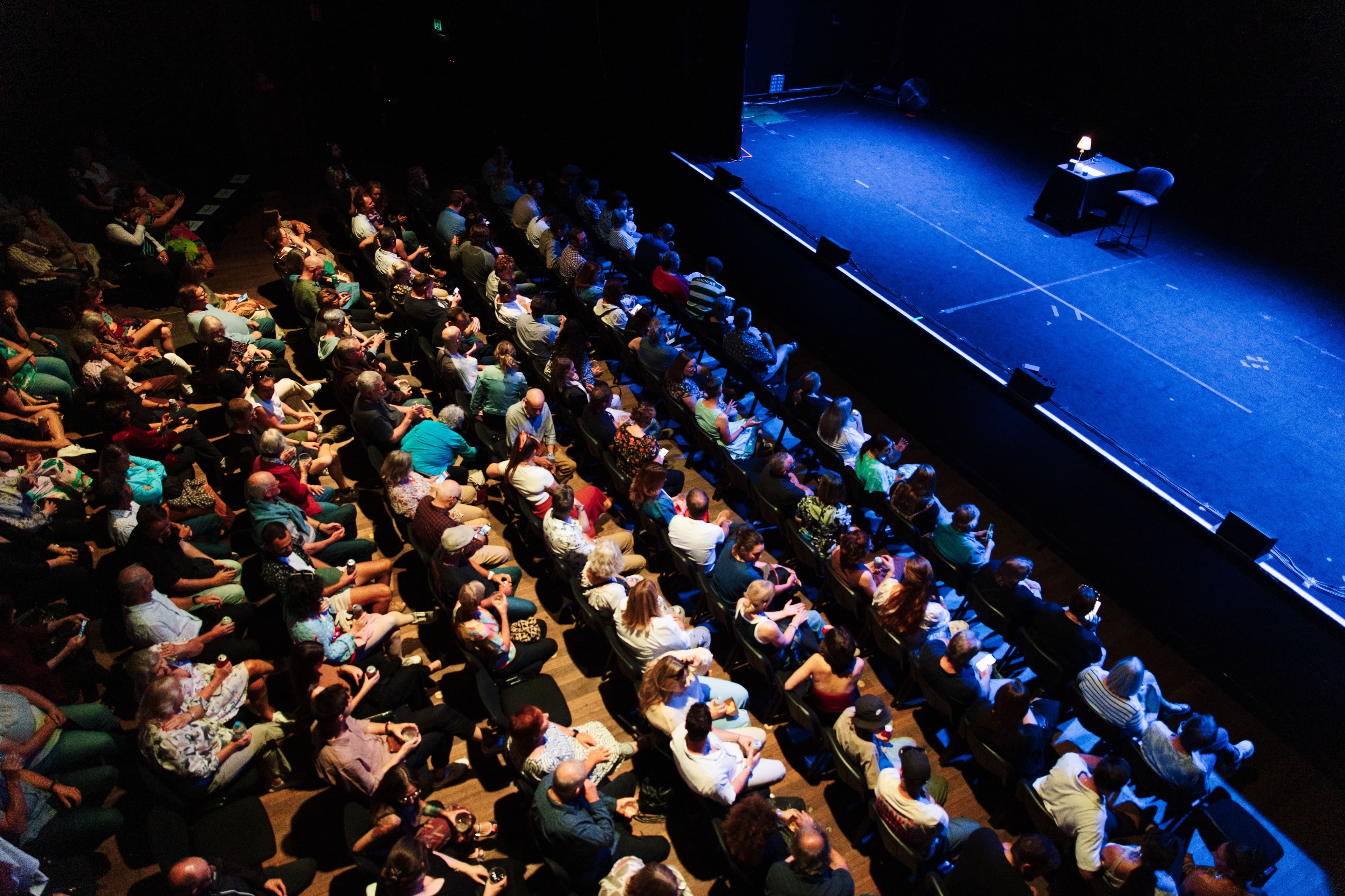 Aerial view of a packed theater audience facing an empty stage with a chair and table. The scene conveys anticipation and excitement.
