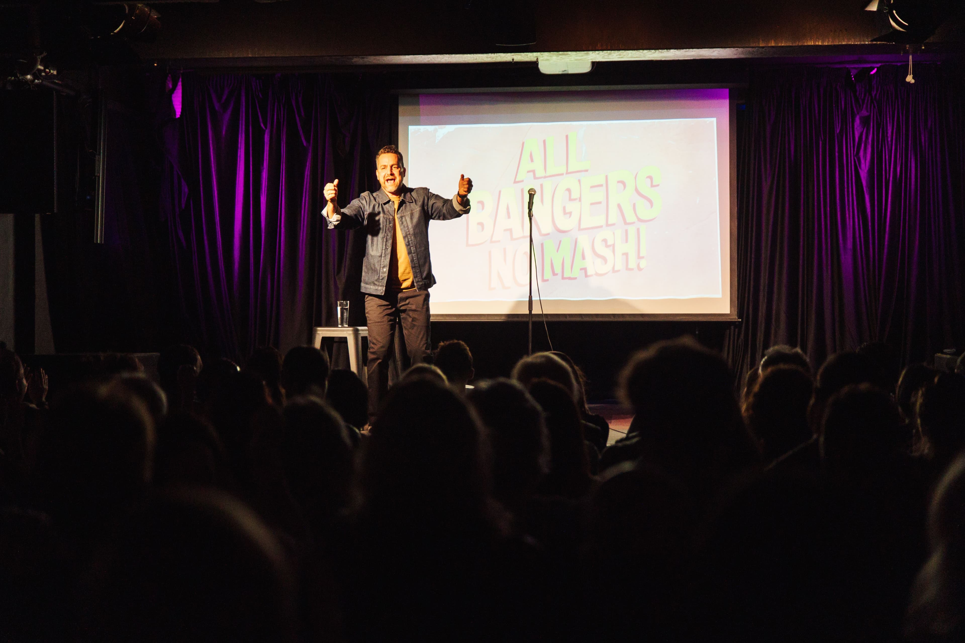 A comedian performs energetically on stage in front of a crowd, gesturing with both hands. Behind him, a screen reads "All Bangers No Mash!"