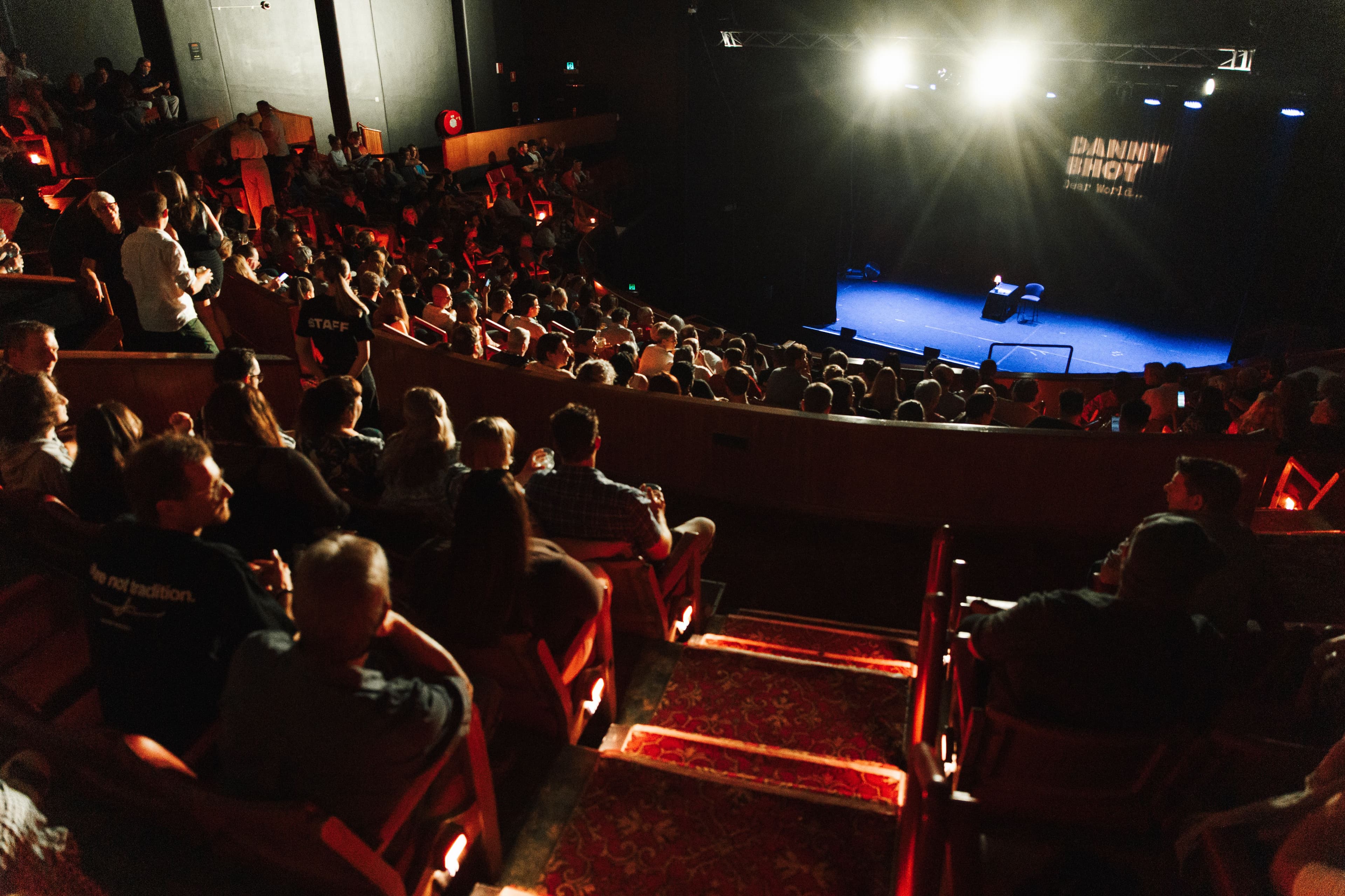 A packed theatre with rows of seated guests facing a brightly lit stage set up for a performance.