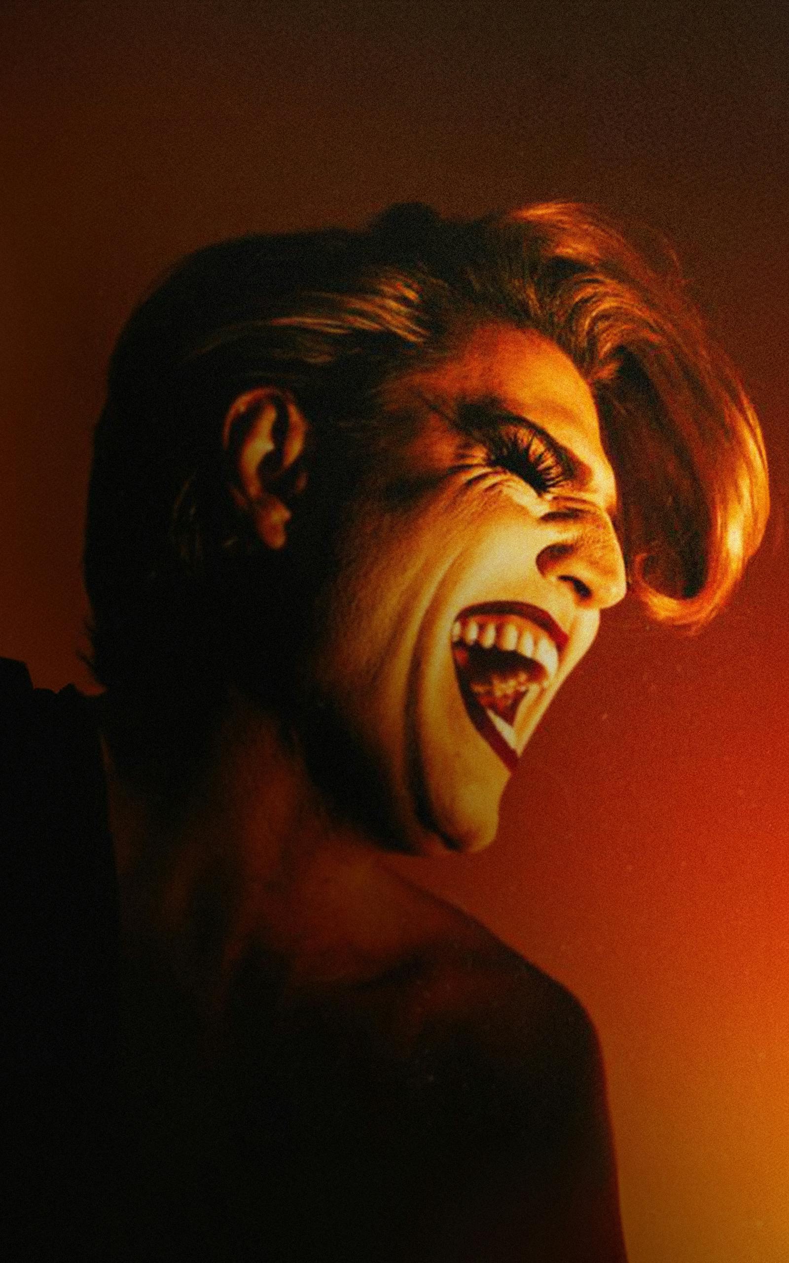 Comedian Reuben Kaye, wearing high contrast makeup and hair styled in a bonny quiff, laughs against a dramatic orange backdrop.